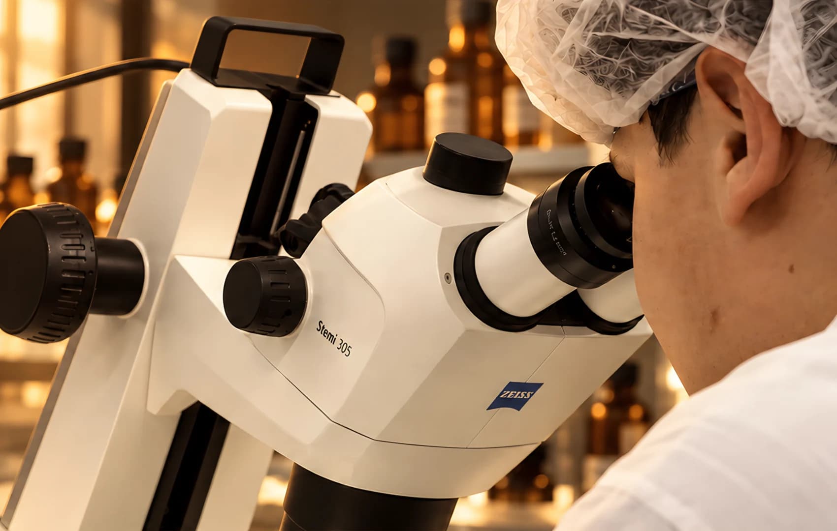 Une personne en charlotte observe un échantillon au microscope, devant des flacons ambrés.