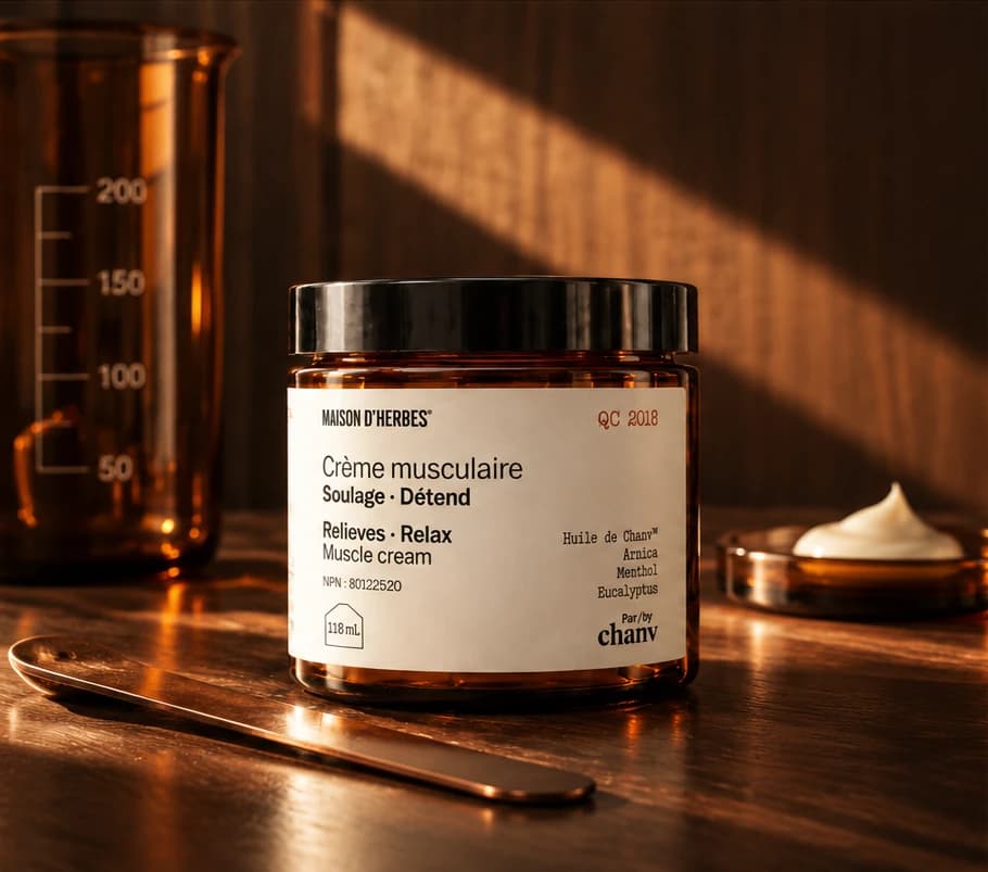 Un pot de Crème musculaire Maison d’Herbes sur une table de bois, dans un rai de lumière chaude, à côté d’un bécher ambré et d’une coupelle de crème