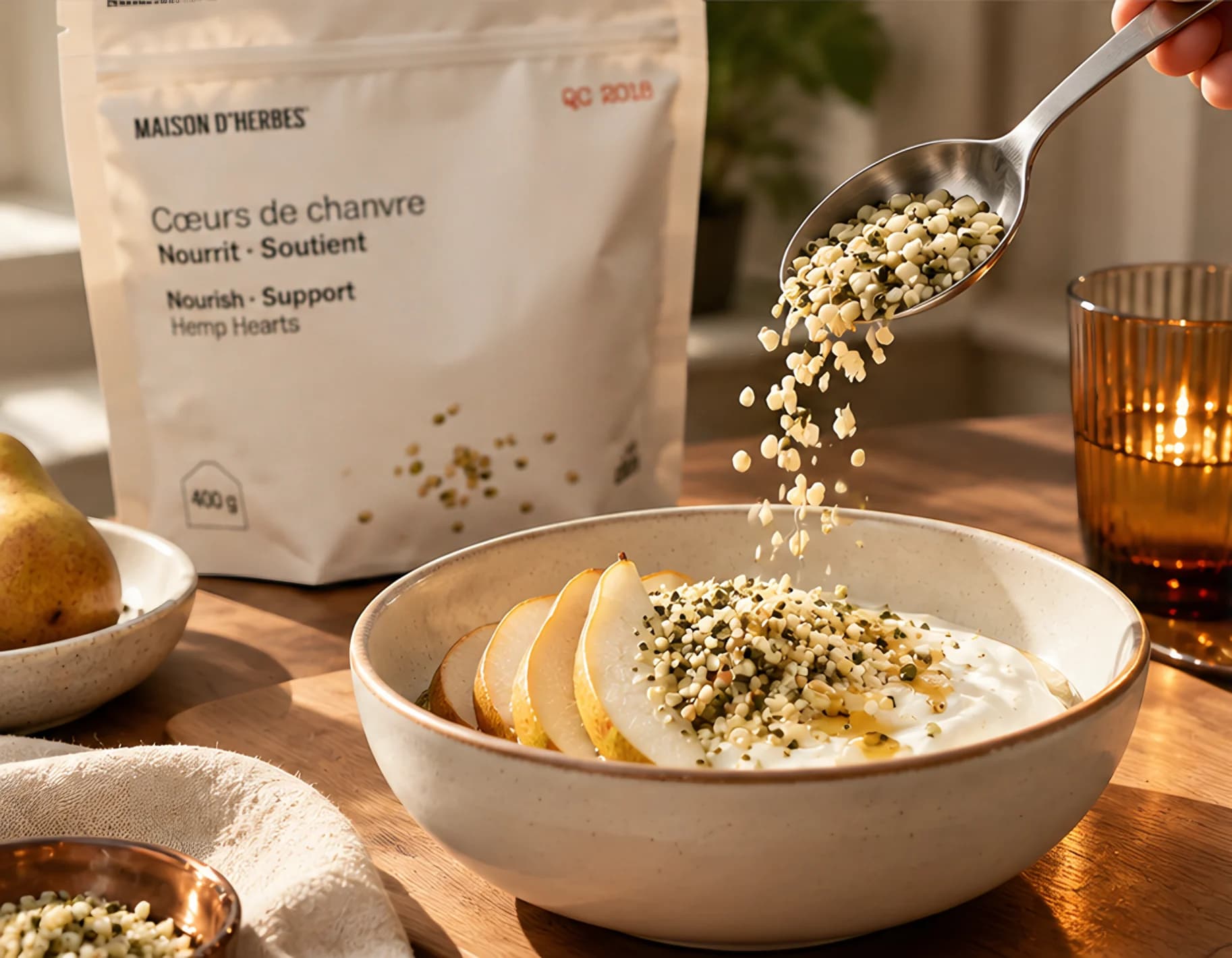 A spoon pouring hemp hearts over a bowl of yogurt and pears, in front of a Maison d’Herbes Hemp Hearts pouch and an amber glass
