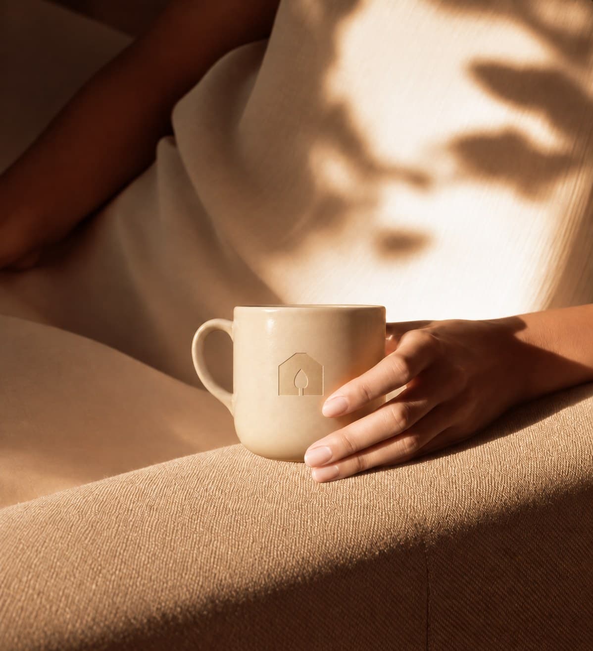 Main posée sur une tasse crème gravée du mot-symbole Maison d’Herbes, ombres de feuillage sur un drapé beige