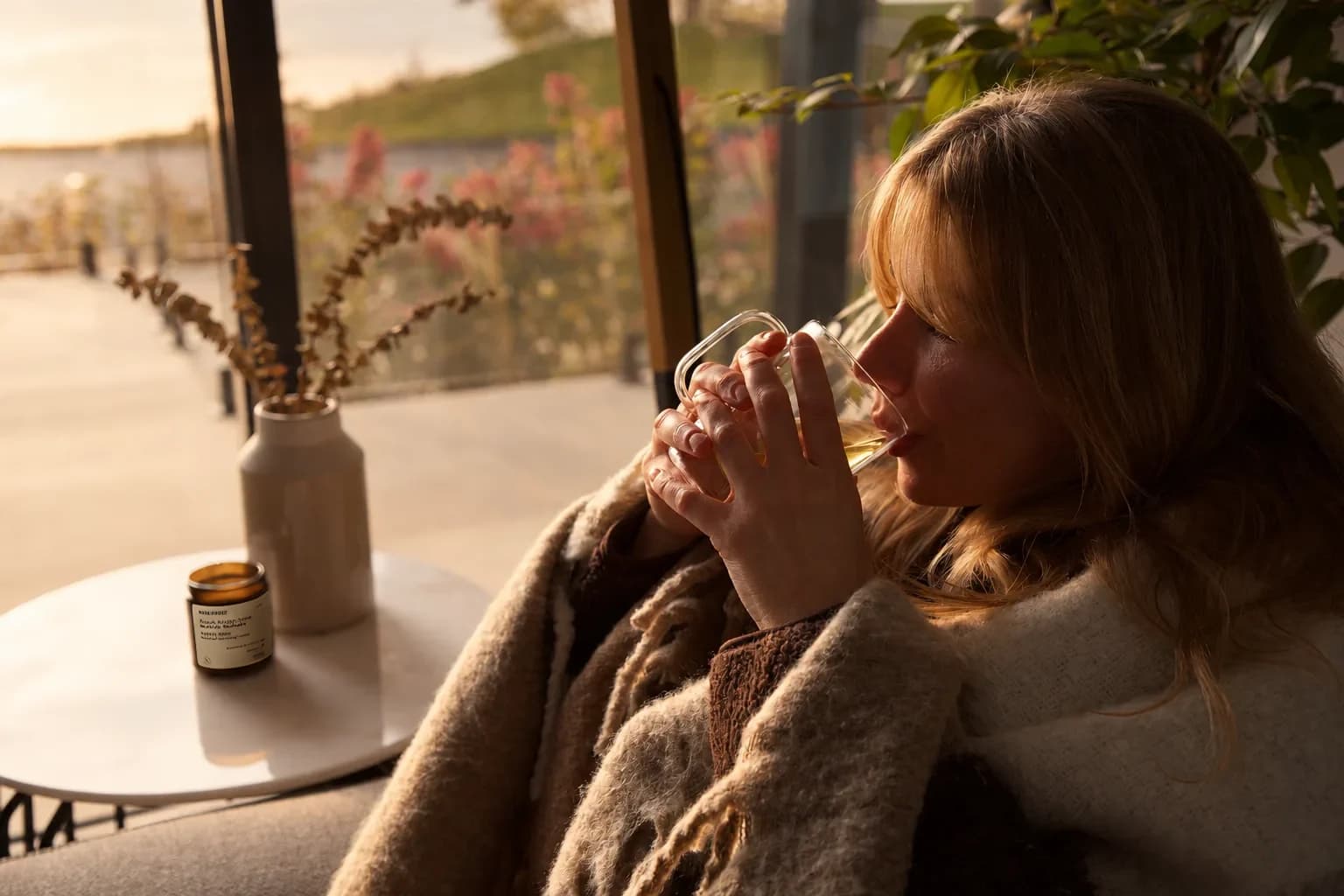 Une femme enveloppée d’une couverture boit une infusion dans une tasse de verre, devant une baie vitrée au soleil couchant ; un pot ambré Maison d’Herbes est posé sur la table