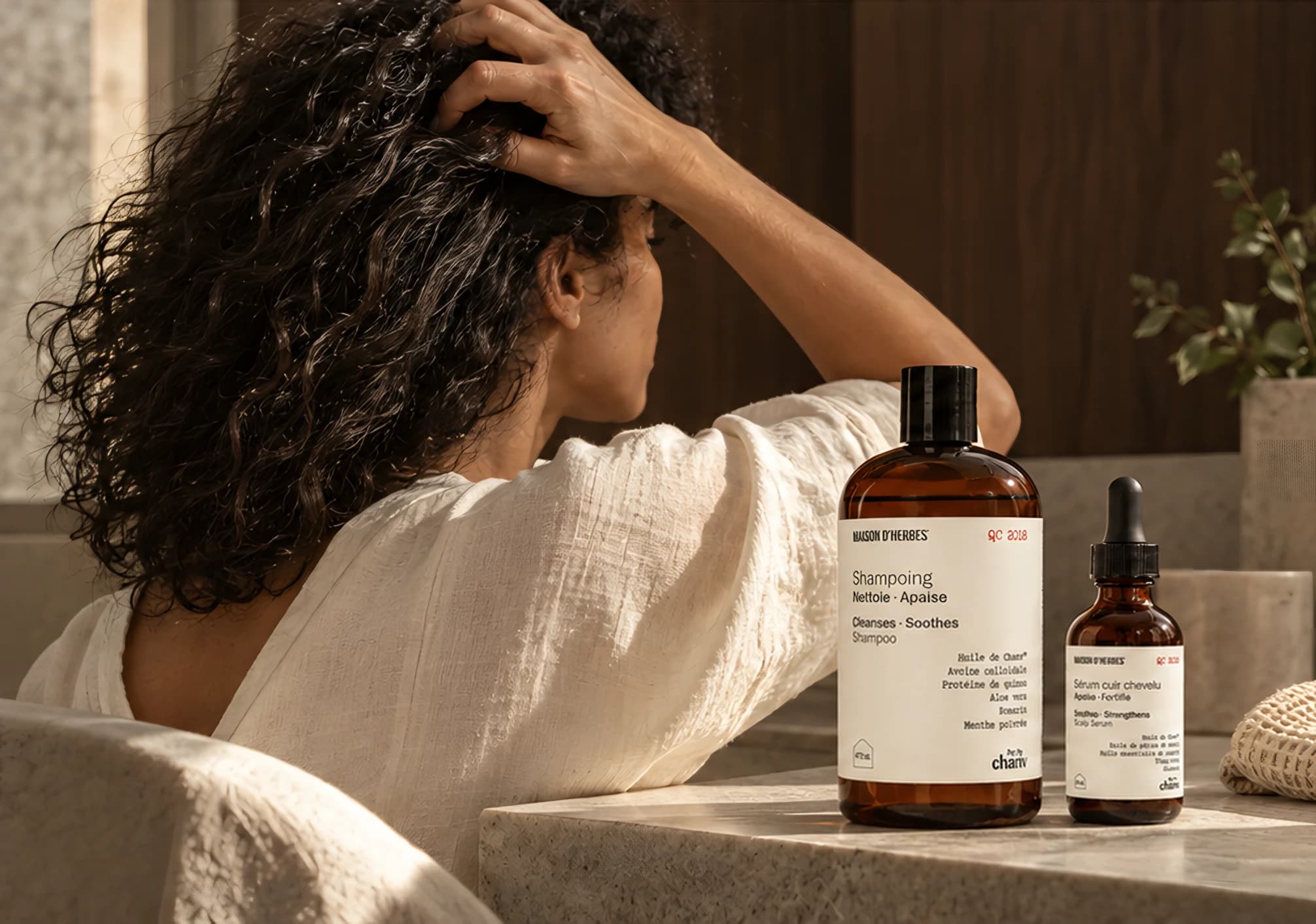 Femme de dos, la main dans ses cheveux bouclés, à côté du Shampoing et du Sérum cuir chevelu Maison d’Herbes posés sur un comptoir de pierre