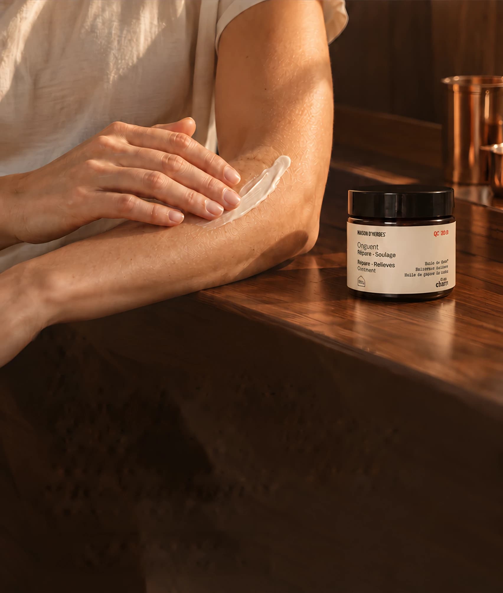 A hand smoothing Maison d’Herbes Repair-Relief Ointment onto a forearm, the jar on a wooden counter