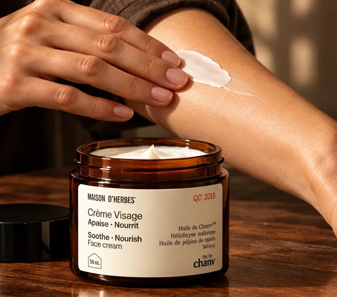 Une main étale la Crème Visage Maison d’Herbes sur un avant-bras, le pot ouvert sur une table de bois