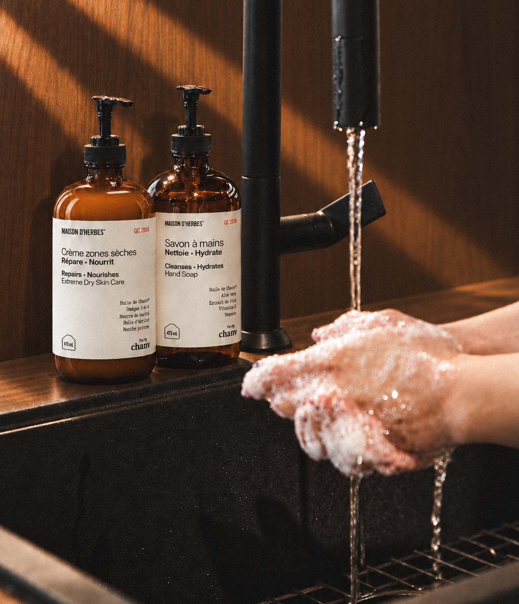La Crème zones sèches et le Savon à mains Maison d’Herbes au bord d’un lavabo, des mains savonnées sous le robinet.